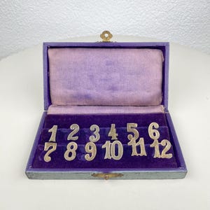 May include: An open, antique, purple velvet lined box with twelve silver-colored numbers. The numbers are arranged in two rows of six. The box has a gold-colored clasp and a decorative top.