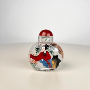 Op de afbeelding: Een helder glazen snuifflesje met een rond lichaam en een effen rode dop. De buitenkant is voorzien van een handgeschilderde illustratie van een figuur in een rood en blauw gewaad, geplaatst bij groen gebladerte en een donkere rotsformatie.