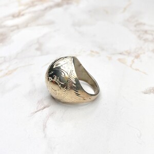 Op de afbeelding: Een zilverkleurige ring met een gegraveerd bloemmotief. De ring heeft een afgeronde bovenkant en een dikke band. Het gegraveerde patroon bevat bladeren en andere botanische elementen. De ring ligt op een wit-grijze marmeren ondergrond.