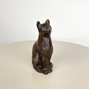 Può includere: Una piccola statuetta di gatto color marrone scuro con una superficie ruvida e testurizzata in posizione seduta. La scultura presenta riflessi color bronzo, orecchie a punta e una coda arrotolata attorno alla base, rivolta verso l'alto.