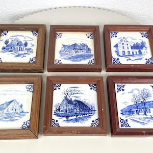 May include: Six framed ceramic tiles with blue and white illustrations of houses and buildings. The tiles are set in brown wooden frames. Each tile has a different house scene, with decorative blue corner accents.
