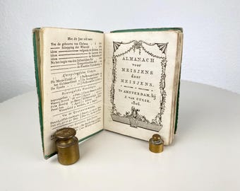 Op de afbeelding: Een open antiek boek met groene binding, getiteld "ALMANACH voor MEISJENS" (Almanak voor meisjes) door J. van Gulik, 1806. Twee kleine messing gewichten zijn bij het boek geplaatst. De pagina's van het boek tonen tekst in het Nederlands.