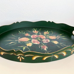 Könnte beinhalten: Ein ovales, dunkelgrünes Serviertablett mit einem zentralen, handgemalten Blumenmotiv aus rosa und pfirsichfarbenen Rosen und grünen Blättern. Das Tablett hat einen gewellten Rand mit goldfarbenen Akzenten und zwei integrierte Griffe.