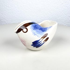 Può includere: Una zuccheriera a forma di uccello in ceramica con base bianca, ornata da pennellate blu, marroni e rosa. La zuccheriera ha un beccuccio e un design simile a una maniglia, ideale per servire panna o salse. Il design dell'uccello è stravagante e decorativo.