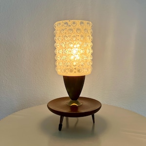 Op de afbeelding: Een vintage tafellamp met een cilindrische, getextureerde glazen kap. De kap is verlicht en geeft een warme gloed. De lamp staat op een donkerbruine houten voet met een kleine, ronde tafel en drie korte poten. De metalen accenten geven elegantie.