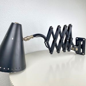 May include: Black, adjustable wall-mounted lamp with a conical shade. The shade has star-shaped cutouts near the bottom. The lamp features a concertina arm and a rectangular wall mount. The lamp's metal components are black and silver.
