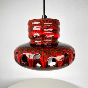 May include: A ceramic pendant light fixture featuring a glossy, lava-style glaze in vibrant red and deep brown tones. The rounded, wide-bottomed shade is decorated with a series of circular cutouts around the lower edge and a black cord.