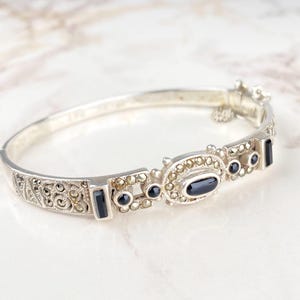 Op de afbeelding: Een zilverkleurige scharnierende armband met een sierlijk ontwerp. De armband heeft een ovale zwarte steen in het midden, omringd door kleinere zwarte stenen en markasiet accenten. De sluiting is ook versierd met kleine stenen.