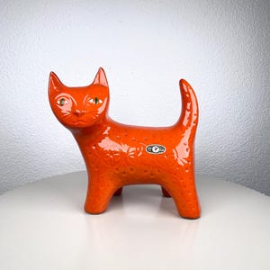 Könnte beinhalten: Orangefarbene Keramik-Katzenfigur mit strukturierter Oberfläche und grünen Augen. Die Katze hat einen gebogenen Schwanz und steht auf vier Beinen. Auf der Seite befindet sich ein kleiner Aufkleber.
