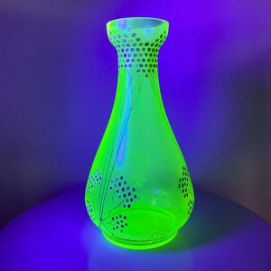 May include: A vibrant green glass vase with a flared base and a narrow neck. The vase has a decorative pattern of small circles and stylized floral designs. The vase is illuminated, creating a bright and eye-catching display. The vase is approximately 25 cm tall.