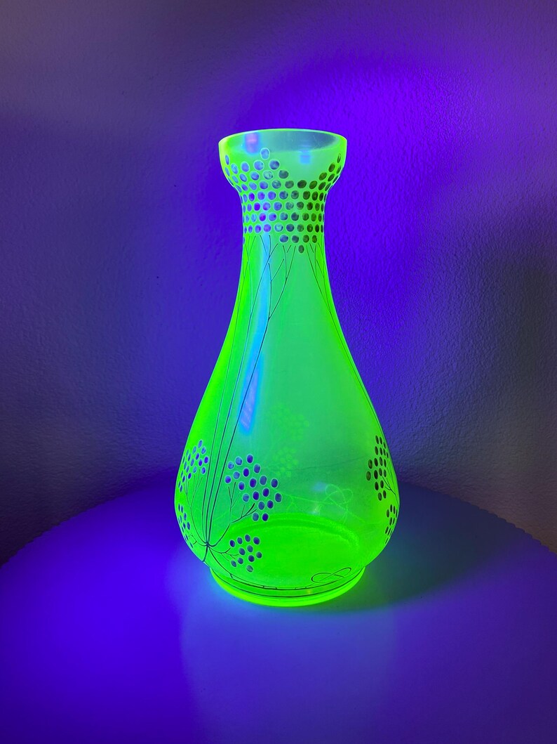 May include: A vibrant green glass vase with a flared base and a narrow neck. The vase has a decorative pattern of small circles and stylized floral designs. The vase is illuminated, creating a bright and eye-catching display. The vase is approximately 25 cm tall.