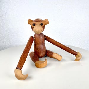 May include: A wooden monkey figurine with articulated limbs. The monkey has a brown body, light brown ears, and a light brown face. The arms and legs are jointed, allowing for posing. The monkey is sitting on a white surface.