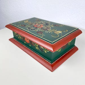 Op de afbeelding: Een rechthoekige houten doos met een scharnierend deksel. De doos is donkergroen geschilderd met een rode rand. Het deksel is versierd met een bloemenontwerp in rood, geel en wit. De doos staat op een wit oppervlak.