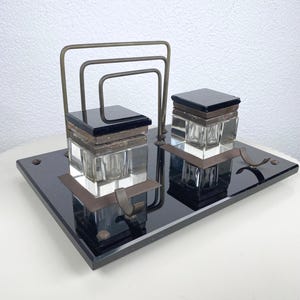 Pode incluir: Um conjunto de secretária antigo com uma base retangular preta, dois tinteiros de vidro com tampas quadradas pretas e um porta-cartas de metal. Os tinteiros são transparentes e o metal tem um acabamento bronze.