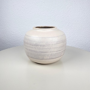 Op de afbeelding: Een ronde, crèmekleurige keramische vaas met horizontale grijze strepen. De vaas heeft een brede, open bovenkant en een licht gestructureerd oppervlak. De binnenkant van de vaas is donkerbruin. De vaas staat op een wit oppervlak.