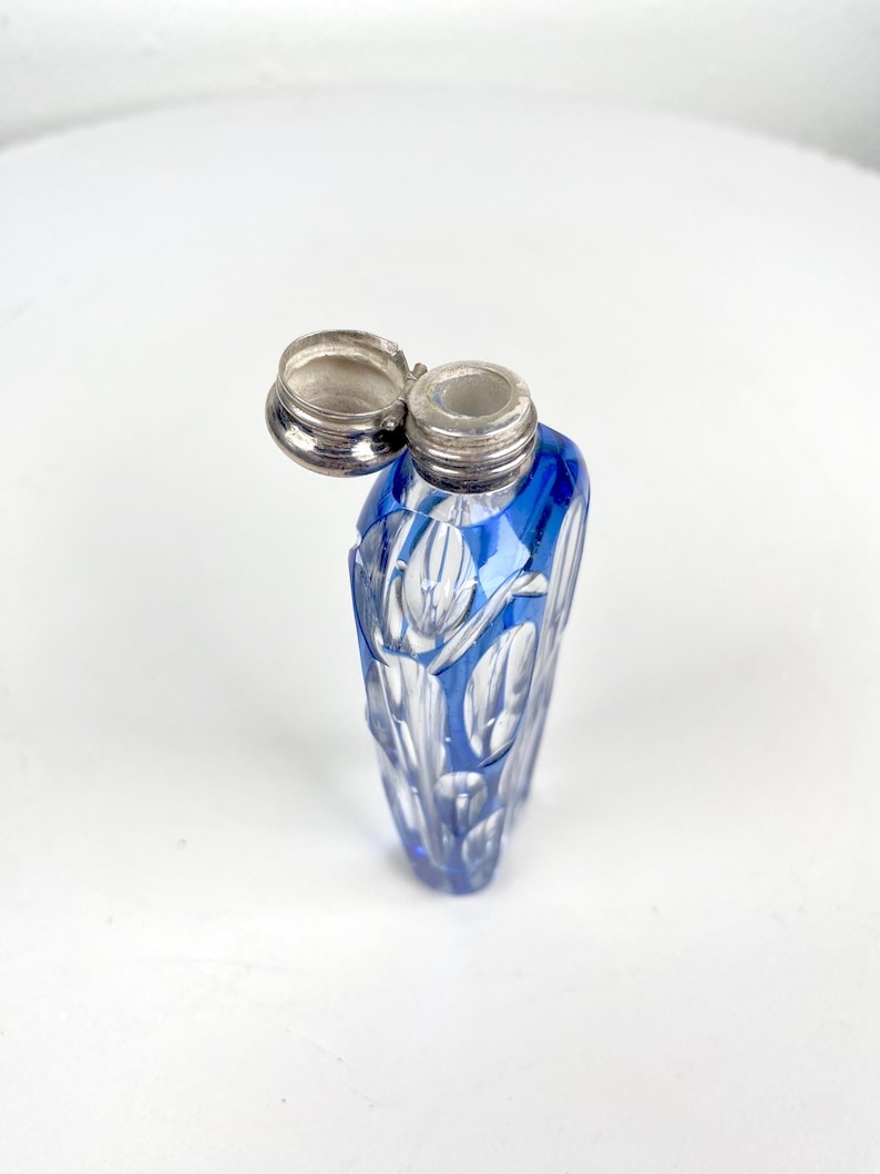 May include: A blue glass perfume bottle with a silver lid. The bottle has a cut glass design with a floral pattern. The lid is open, revealing the bottle's opening. The bottle is approximately 4 inches tall.