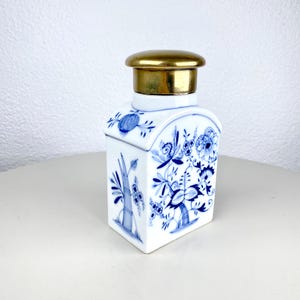 Op de afbeelding: Een witte porseleinen theebus met blauwe bloempatronen en een goudkleurig deksel. De bus heeft een rechthoekige vorm met afgeronde randen en is versierd met gedetailleerde blauwe bloemmotieven. Het deksel heeft een glanzende goudkleur.