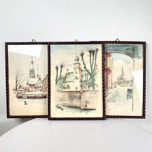 May include: Three framed watercolor paintings of European architecture. The artwork features detailed depictions of buildings, churches, and waterways, with a muted color palette. The frames are dark brown with a textured design, adding a vintage aesthetic.