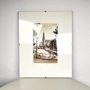 Op de afbeelding: Een ingelijste sepia-kleurige prent met een kanaalscène, fietsen geparkeerd langs een reling, een hoge kerktoren op de achtergrond en bomen langs het water. Het kunstwerk zit in een glazen lijst met metalen clips aan de randen.