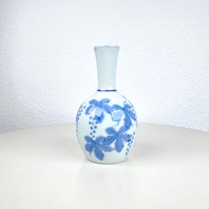 Może przedstawiać: Mały, biały ceramiczny wazon z wąską szyjką i zaokrąglonym korpusem. Wazon jest ozdobiony niebieskim wzorem liści winogron i kiści winogron. Wazon ma około 15 cm wysokości.