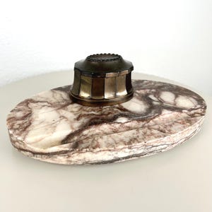 Pode incluir: Um tinteiro antigo com tampa de latão e base de mármore. O tinteiro tem um design geométrico e assenta numa bandeja oval de mármore com veios castanhos e brancos. Material de escritório vintage.