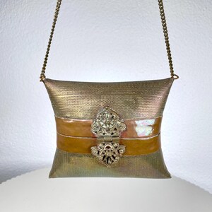 Op de afbeelding: Een vintage-stijl, metallic gouden handtas met een kettingriem. De tas heeft een decoratieve sluiting en een horizontale band van iriserend materiaal. De body van de tas heeft een mesh-achtige textuur en het algehele ontwerp is sierlijk en elegant.