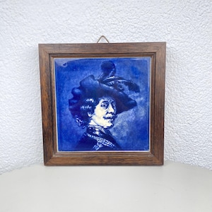 Op de afbeelding: Een ingelijste blauw-witte keramische tegel met een portret van een man met een grote hoed. Het kunstwerk is geplaatst in een donkerbruine houten lijst, wat een klassiek en decoratief stuk creëert.