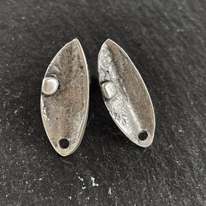 May include: A pair of silver earrings shaped like leaves with a small round bead on each. The earrings have a textured, rustic look.