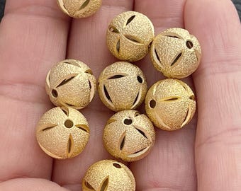 May include: A collection of spherical gold-toned beads featuring a textured, sandblasted finish. Each bead is designed with a series of symmetrical, curved cutouts that reveal a smooth interior surface, creating a distinct pattern across the round shape.