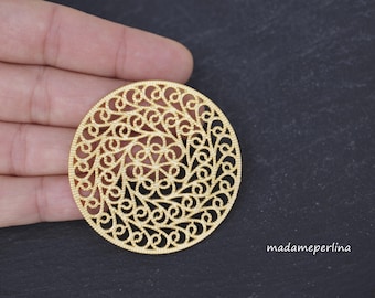 May include: A circular gold-toned metal filigree component featuring an intricate, repeating swirl pattern radiating from a central floral-like motif. The piece has a textured surface and is displayed against a dark background with the text madameperlina.