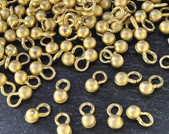May include: A collection of small, matte gold-toned metal ball buttons, each featuring a smooth spherical head attached to a circular loop shank. These individual fasteners are scattered across the frame, showcasing a uniform metallic finish.