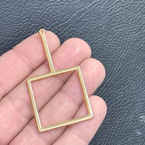 May include: A gold-toned metal pendant in the shape of an open square frame with a vertical bar extending from the top center, featuring a small circular loop at the very top for attachment, characterized by a smooth, reflective, and polished finish.
