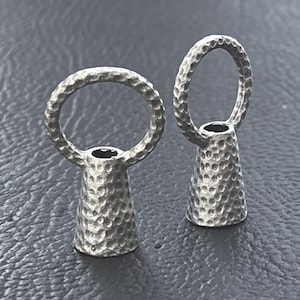Könnte beinhalten: Zwei silberfarbene Kordelenden aus Metall, jeweils mit einer konischen Basis und einem darüber liegenden kreisförmigen Ring. Beide Komponenten weisen eine markante, gehämmerte und genoppte Textur auf der gesamten Oberfläche auf.