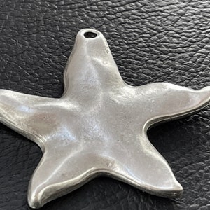 May include: A silver-colored starfish pendant with a loop at the top for a chain. The pendant has a smooth, slightly textured surface and is designed with five rounded points. The starfish is approximately 2 inches wide.