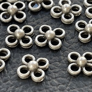 May include: Silver metal flower-shaped charms with five loops. The charms are scattered on a dark grey textured surface.