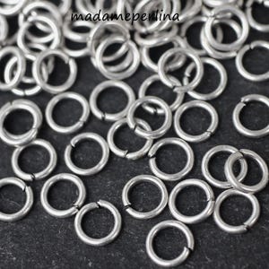 Op de afbeelding: Close-up van een verzameling kleine, zilverkleurige sprongringen. De ringen zijn open en klaar om verbonden te worden. De afbeelding is van bovenaf genomen en toont de ringen verspreid op een donkere ondergrond. De ringen zijn voor het maken van sieraden.