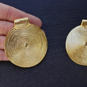 May include: Two gold-colored, circular pendants with a spiral design. Each pendant has a small, rectangular loop at the top. The pendants are displayed on a dark surface.