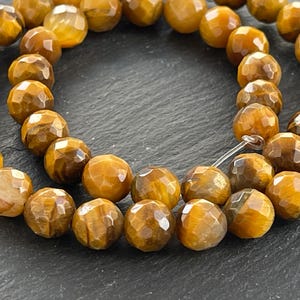 Könnte beinhalten: Strang aus facettierten Tigeraugen-Edelsteinperlen. Die Perlen sind eine Mischung aus goldbraunen und bernsteinfarbenen Farbtönen mit polierter Oberfläche. Die Perlen sind auf einem transparenten Faden aufgereiht, für die Schmuckherstellung.