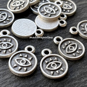 Könnte beinhalten: Eine Gruppe silberfarbener Metall-Anhänger mit einem Auge, das von Sonnenstrahlen umgeben ist. Die Anhänger haben oben ein Loch zum Anbringen an Schmuck oder anderen Bastelarbeiten.