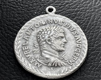 May include: A circular silver-toned metal pendant featuring a relief profile of a bearded man wearing a laurel wreath. The inscription M AVREL ANTONINVS PIVS AVG GERM encircles the portrait, and a small loop is attached to the top edge.