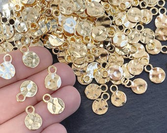 May include: A collection of small, round, gold-colored metal charms featuring a hammered, textured surface. Each charm includes a circular loop at the top for attachment. The charms have a reflective, metallic finish and are scattered in a pile.