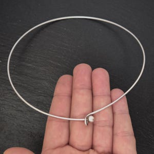 May include: A delicate silver choker necklace with a small, round pearl pendant. The necklace is a simple, elegant design, perfect for everyday wear or special occasions. The choker is held in a hand against a dark background.