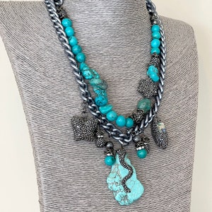 May include: A statement necklace featuring a silver-tone chain, turquoise beads, and various decorative charms. The necklace includes a large turquoise pendant with a snake design and smaller charms with crystal accents. The necklace is displayed on a mannequin.