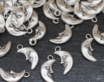 Puede incluir: Un montón de abalorios de metal plateados en forma de medias lunas con una cara en cada luna. Los abalorios tienen un agujero en la parte superior para sujetarlos a joyas u otras manualidades.