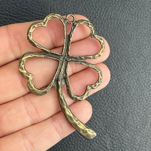 May include: A gold-toned metal four-leaf clover charm with a textured surface. The charm has a loop at the top for attaching to a necklace or bracelet.