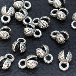 May include: A selection of small, silver-coloured bead caps with a floral filigree design. Each cap has a loop for attaching to jewellery and a hinged, petal-like opening. The caps are scattered across a dark surface.