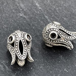May include: Two silver metal bead caps with an intricate design. Each cap has a central opening, encircled by a pattern of small dots and decorative lines. The caps are flower-shaped, with a dark centre.