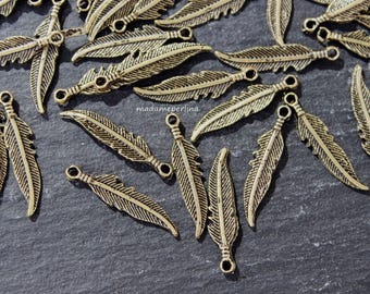 Puede incluir: Una colección de dijes de plumas de metal en tono dorado antiguo, cada uno con una superficie texturizada detallada y un pequeño lazo circular en la parte superior para su fijación. Los dijes están dispersos, mostrando varios ángulos.