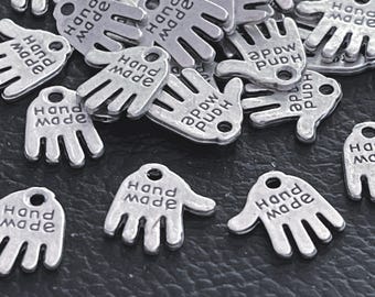 Puede incluir: Una colección de dijes de metal en tono plateado con forma de manos, cada uno con un pequeño orificio en la parte superior para su fijación. La palabra hand made está grabada en relieve en la palma de cada dije.