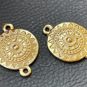 Puede incluir: Dos conectores metálicos circulares de tono dorado con un patrón de rayos de sol en relieve, un punto central, círculos concéntricos y rayos triangulares. Cada pieza incluye dos pequeñas anillas en los lados opuestos para sujeción.